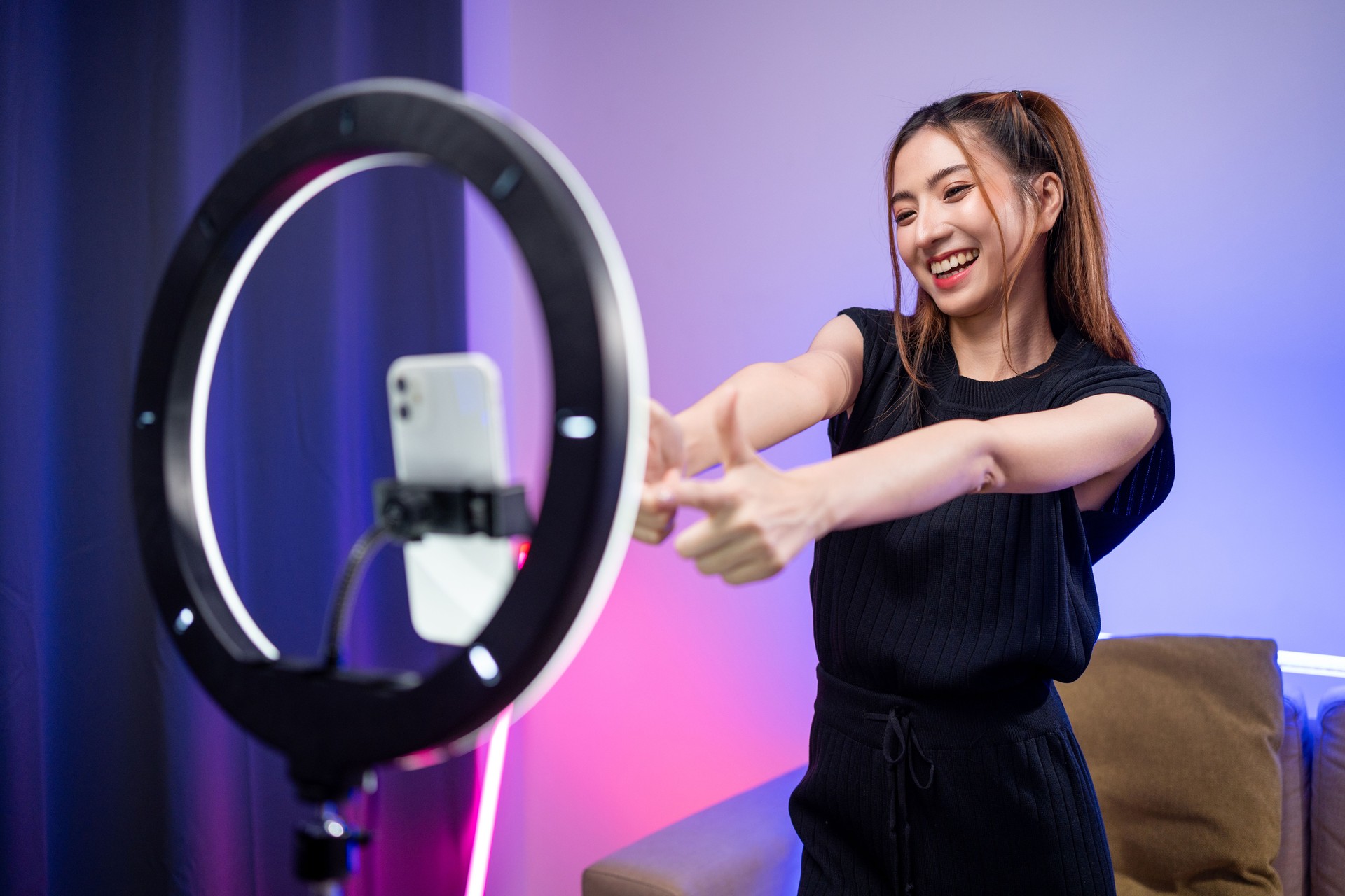 Young trendy influencer asian woman dancing on mobile phone at home in living room with neon light. Creator vlogger talent dancing enjoy hobby content recording show video sharing on social media.